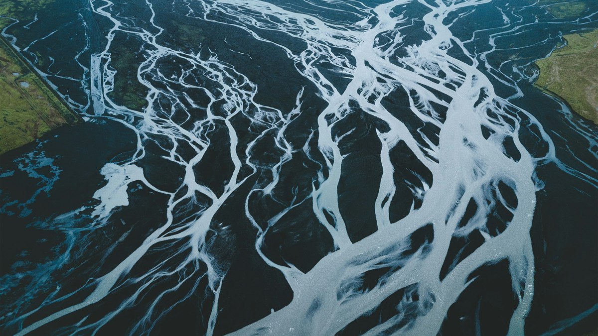 An aerial view of a braided river, where dozens of pale channels split and rejoin across dark gravel flats.  Photo by Rolands Varsbergs on Unsplash