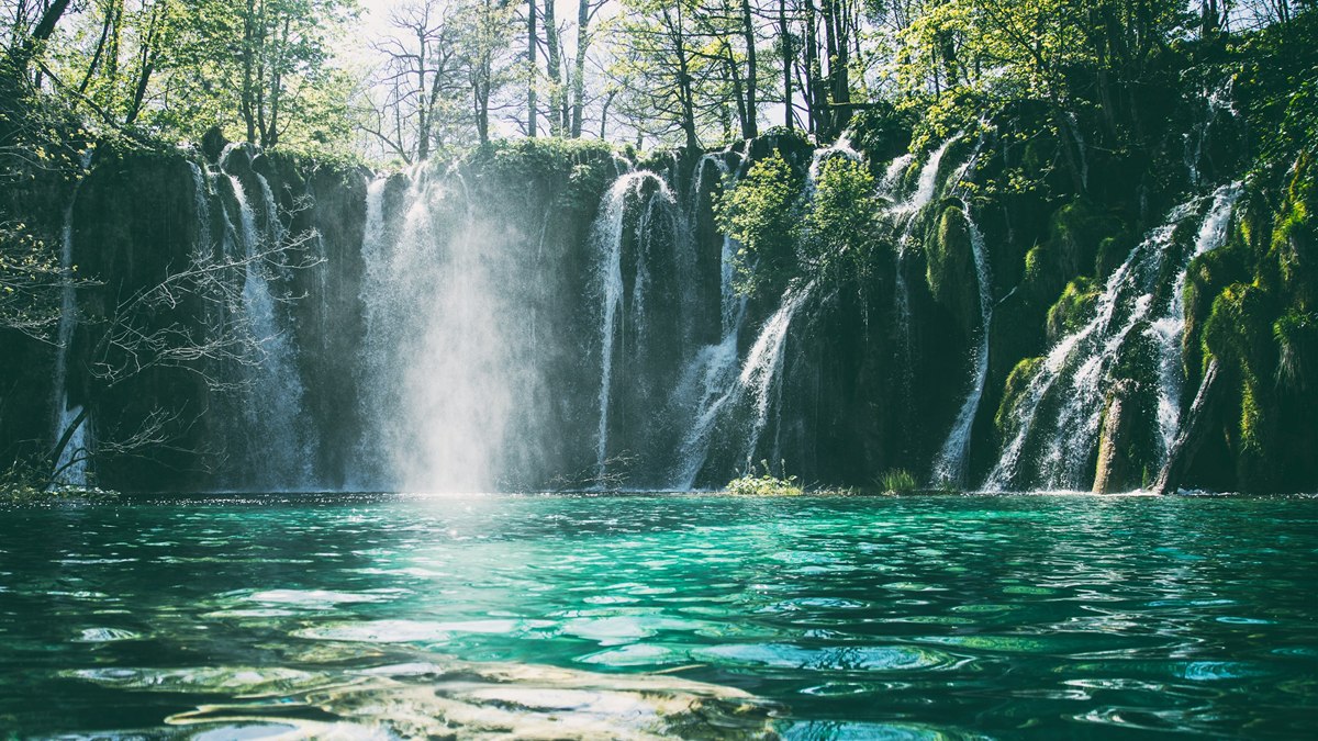A row of many small waterfalls spilling over a mossy, tree-lined ledge into a clear turquoise lake.